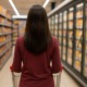 Eine Frau steht mit dem Rücken zur Kamera in einem Supermarkt und schiebt einen Einkaufswagen durch eine Reihe mit Fertigprodukten.