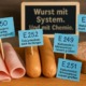 Verarbeitete Fleischprodukte mit blauen Fähnchen: E250, E251, E252, E249 – Hinweise auf Nitrit- und Nitratzusätze; im Hintergrund eine Tafel mit dem Text „Wurst mit System. Und mit Chemie“