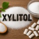 Holzlöffel mit Xylit-Kristallen, Glas mit Birkenzucker, Xylit-Kaugummis und grüne Birkenblätter auf Holztisch arrangiert
