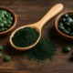 Holztisch mit zwei Schalen Spirulina- und Chlorella-Tabletten sowie einem Holzlöffel voller Spirulina-Pulver